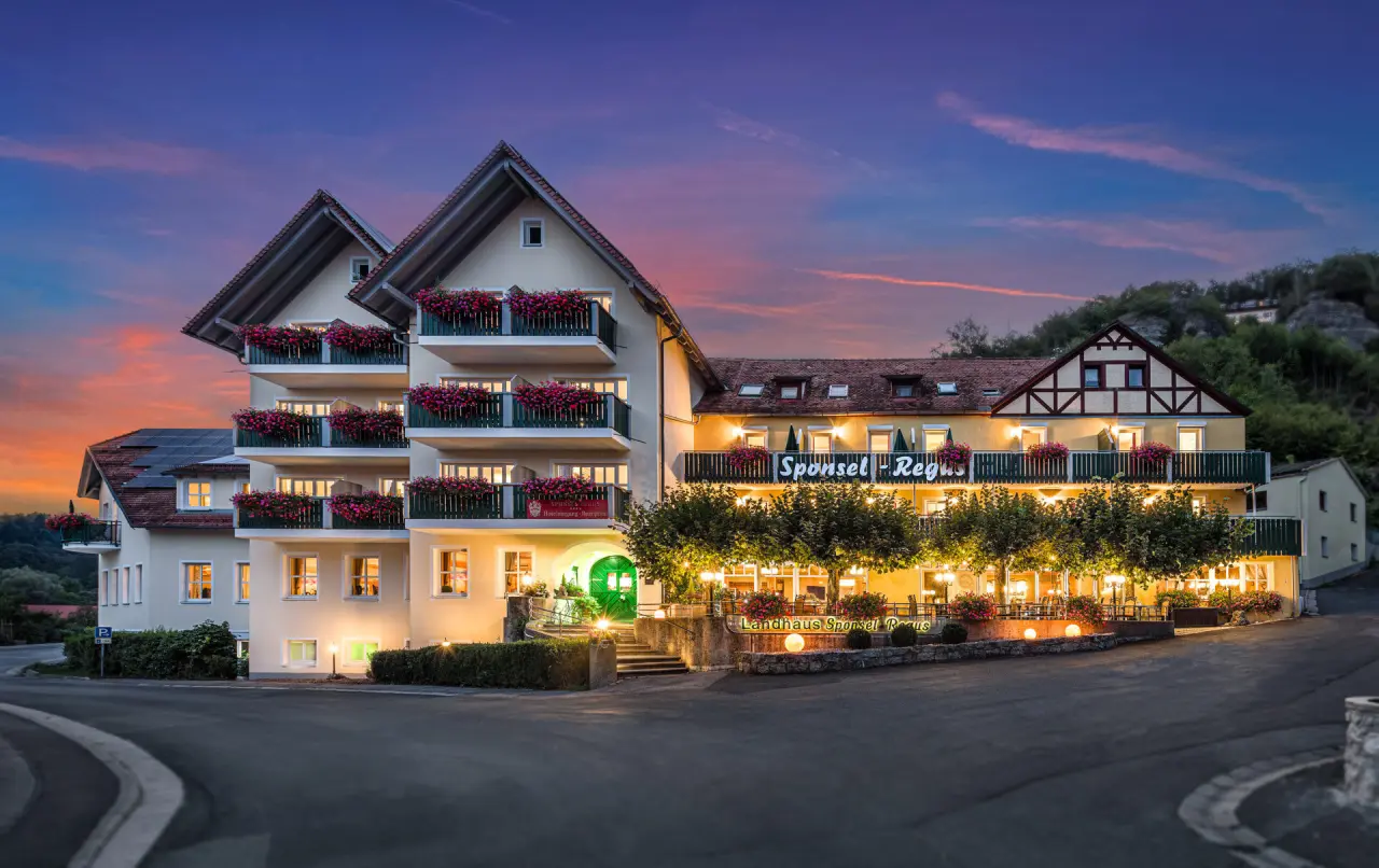 Charmantes Landhaus Sponsel-Regus bei Sonnenuntergang mit beleuchteten Balkonen und blühenden Blumen.