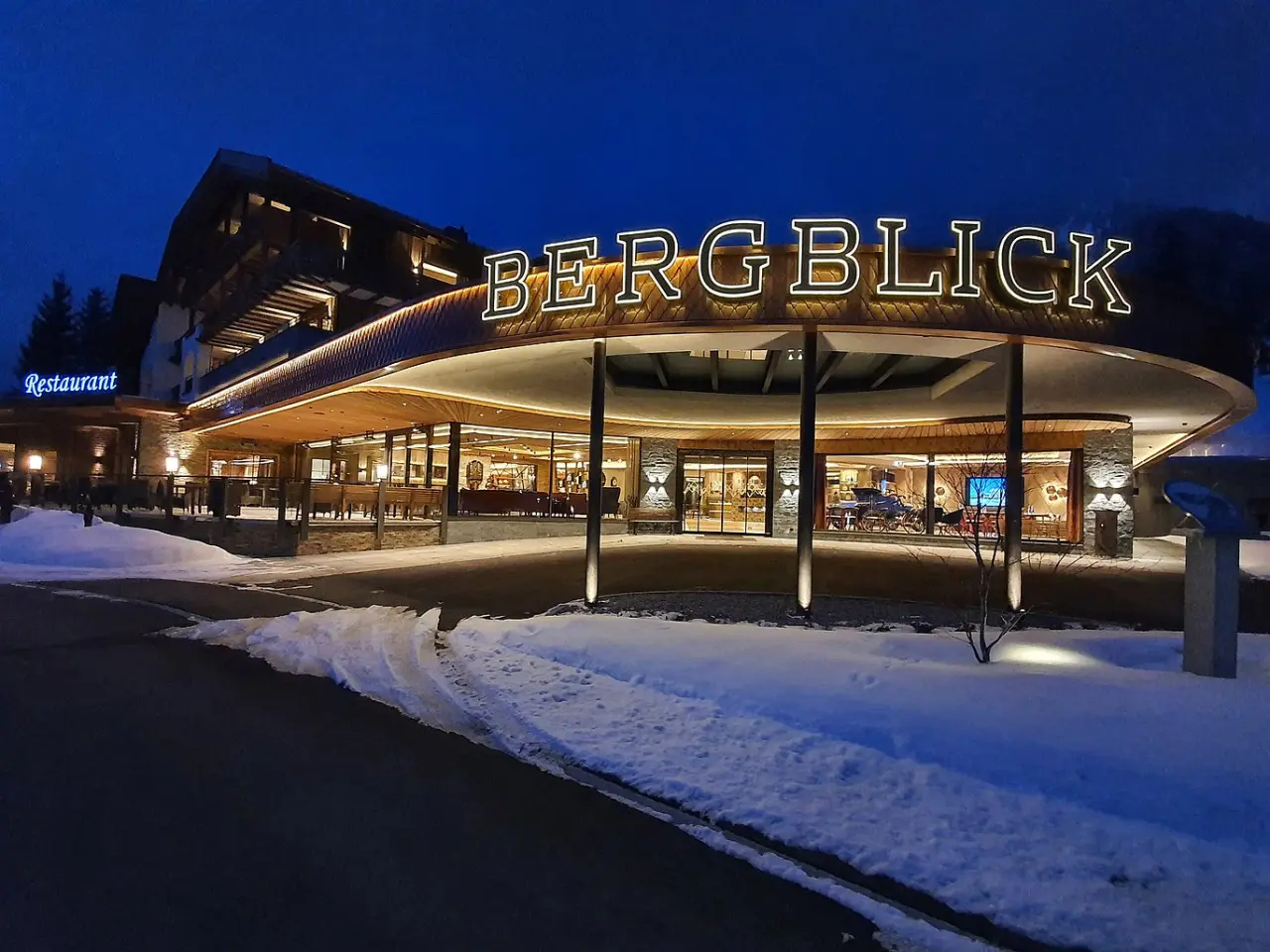 Winterliche Nachtansicht des 5-Sterne Hotel Bergblick mit beleuchtetem Schriftzug und einladendem Eingangsbereich im Schnee.