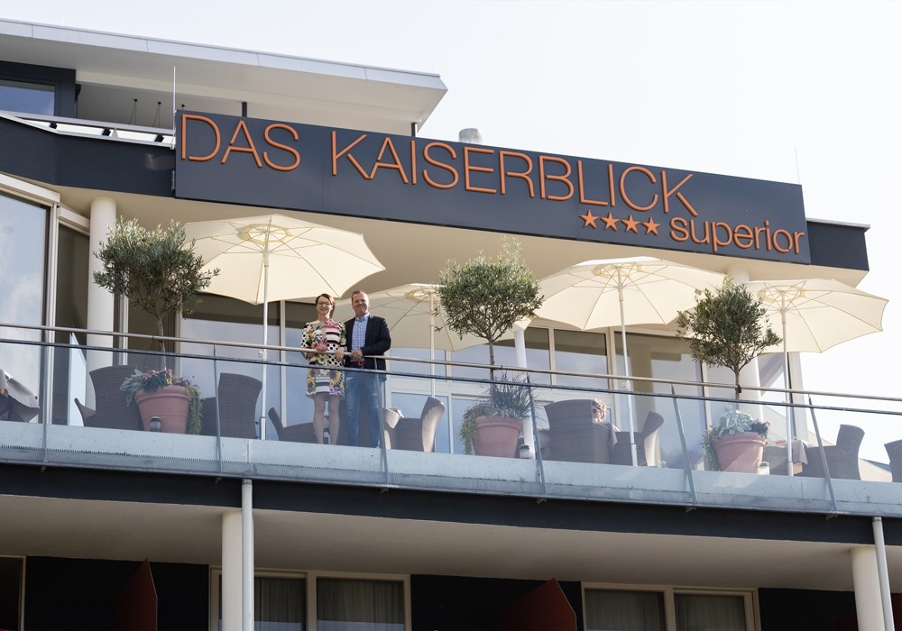 Hotel Kaiserblick in Ellmau – Hotellogo und Balkonbereich mit Sonnenschirmen und Hotelpersonal | Wellnesshotel Tirol