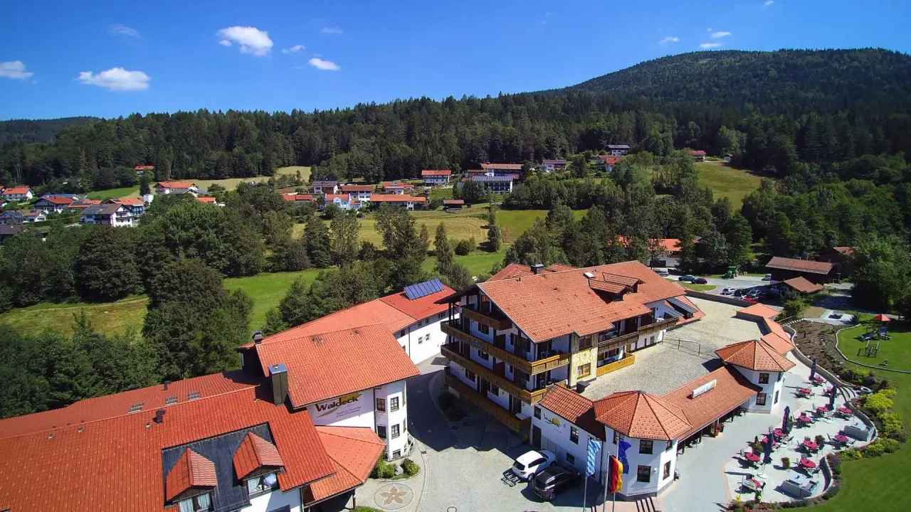 Luftaufnahme des Hotel Waldeck mit roten Dächern, umgeben von grünen Wäldern und Wiesen unter blauem Himmel.