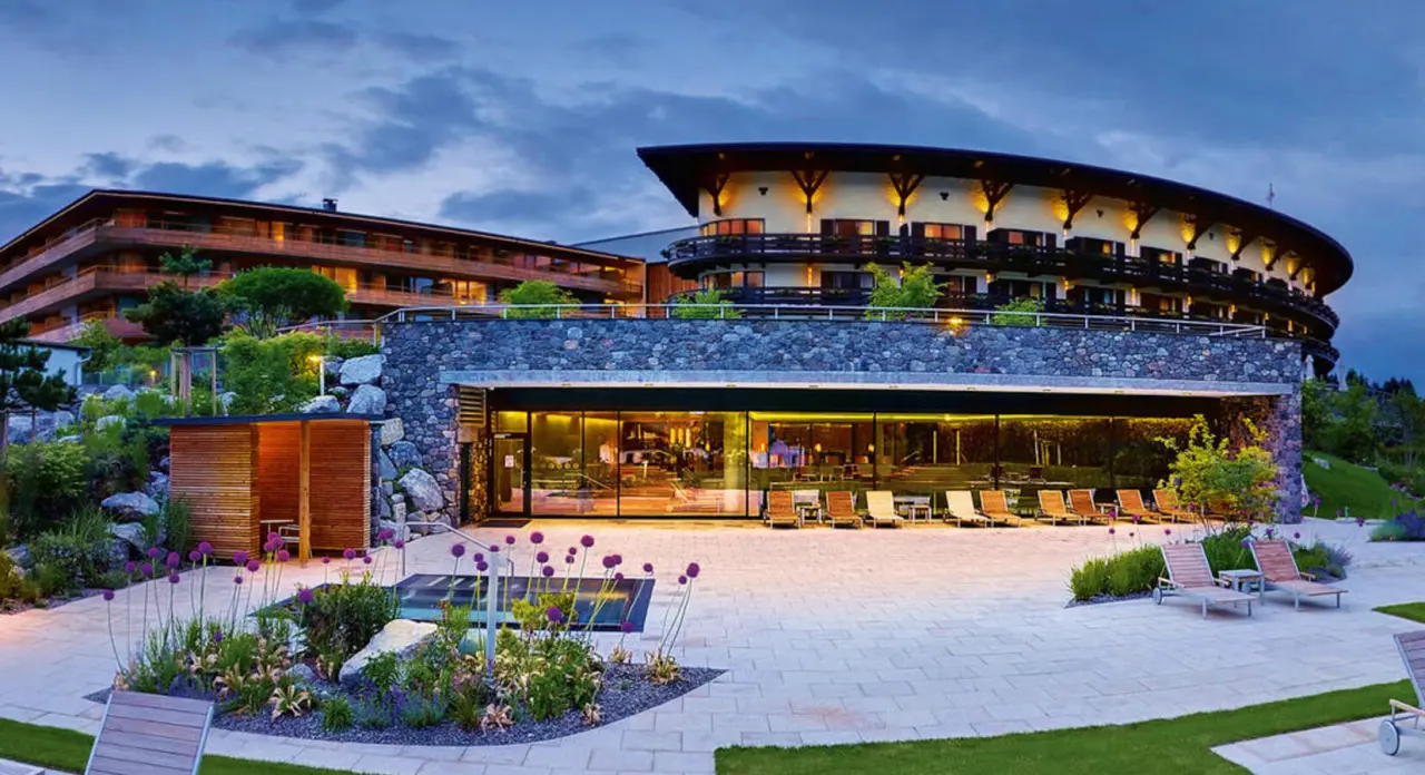 Travel Charme Ifen Hotel bei Abenddämmerung: Moderne Architektur, beleuchtete Fassade und entspannender Aussenbereich mit Liegen.