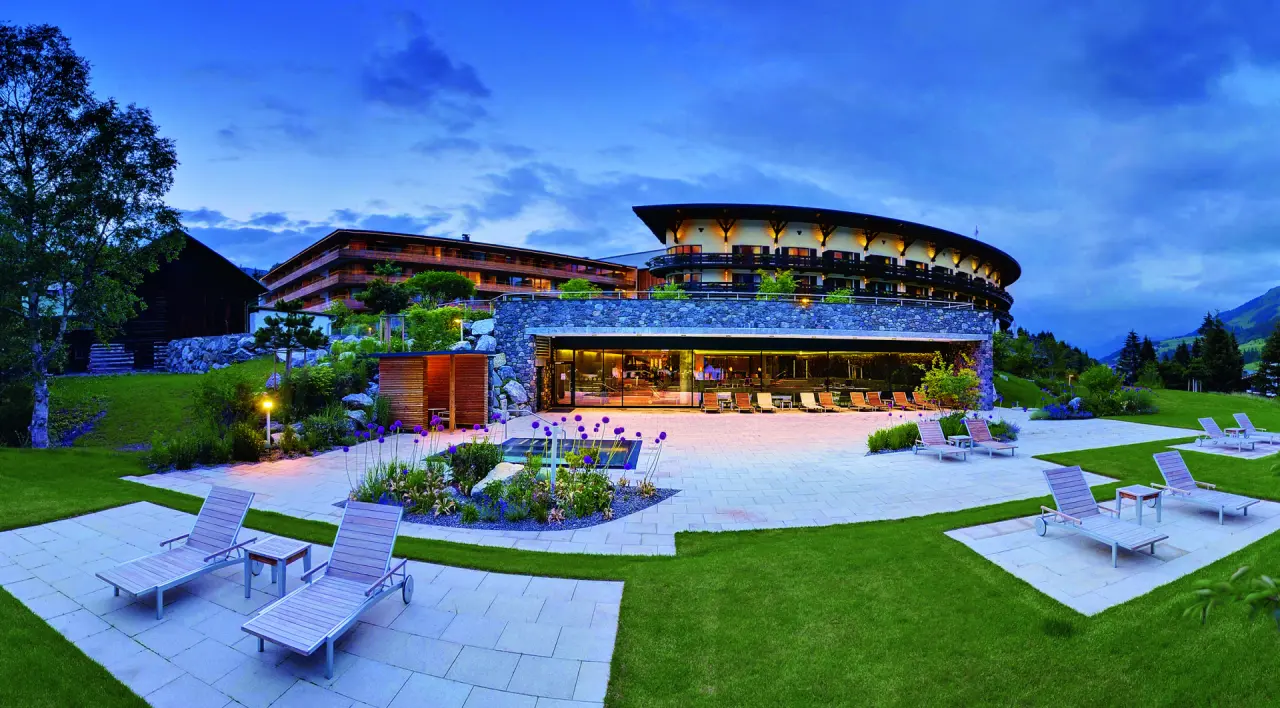 Abendliche Aussenansicht des Travel Charme Ifen Hotel mit beleuchteter Terrasse, Liegestühlen und Bergblick.