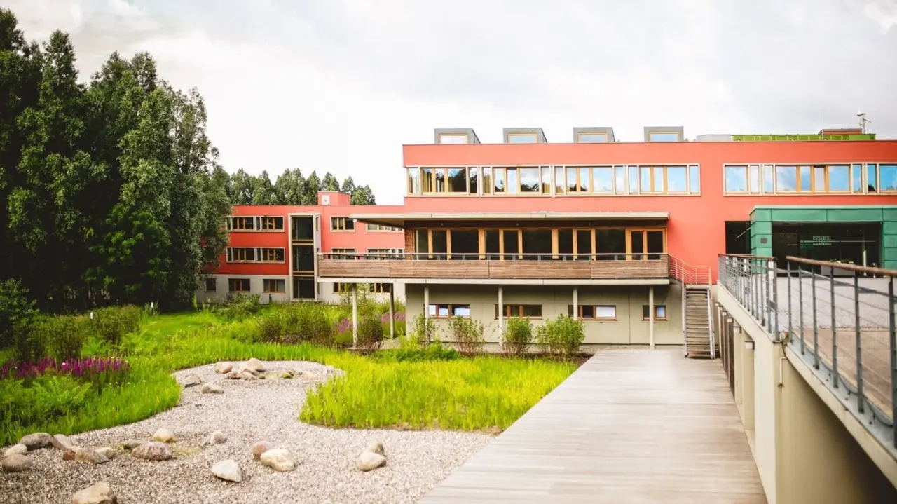 Modernes rotes Hotelgebäude des Ostseehotel Dierhagen mit grüner Gartenanlage und Holzsteg unter bewölktem Himmel.