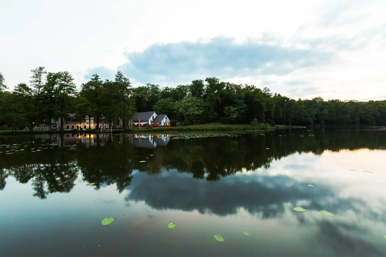 Abendliche Aussenansicht des Gut Klostermühle Alt Madlitz am See, umgeben von Bäumen, mit Spiegelung im Wasser.