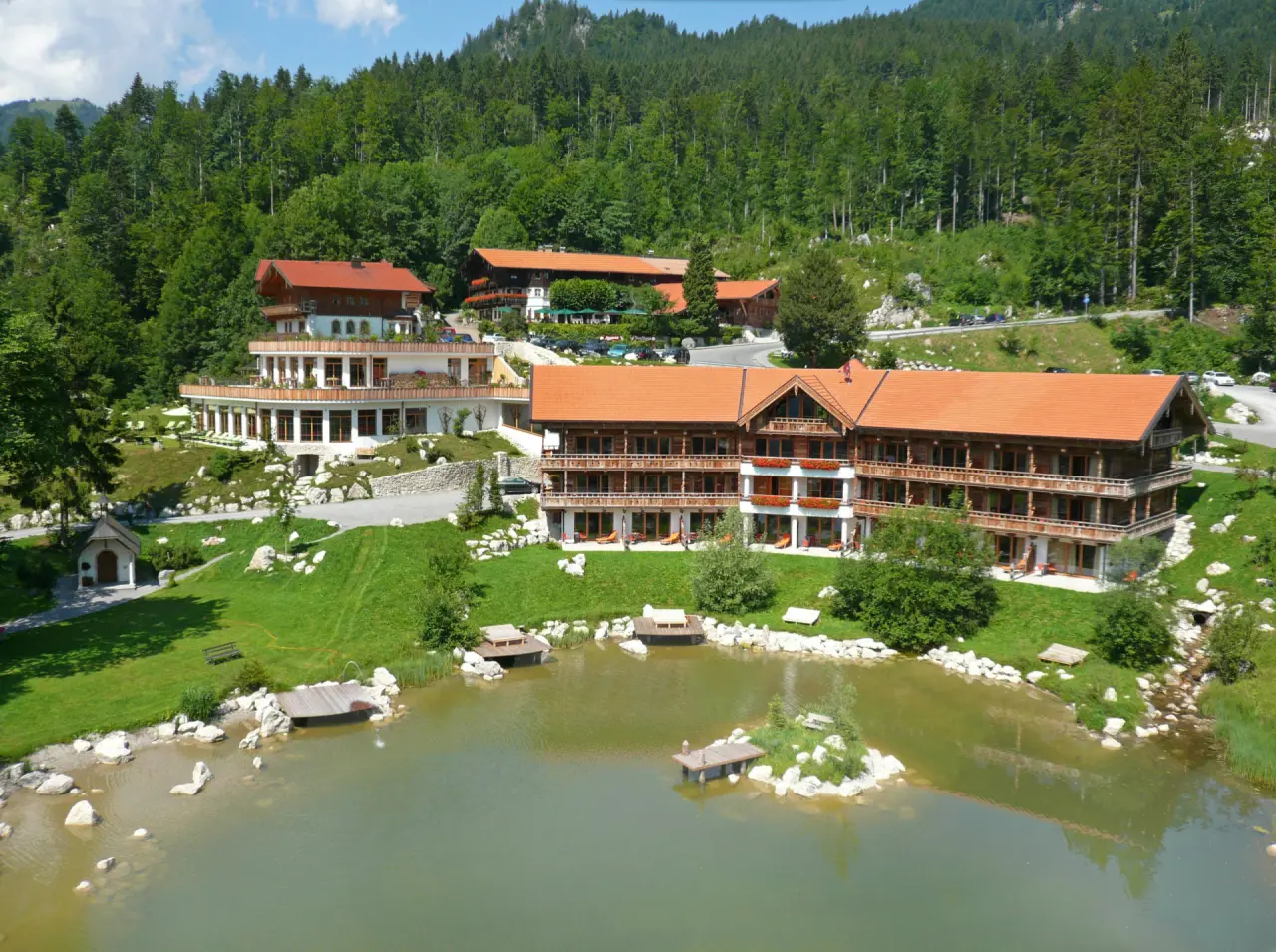 Gesamtansicht des Hotels Feuriger Tatzlwurm mit See und Bergpanorama in den bayerischen Alpen.