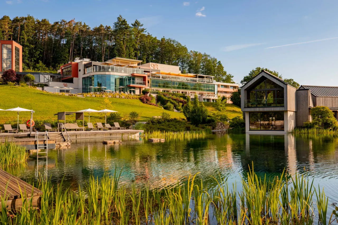 Pfalzblick Wald Spa Resort mit Naturteich, Liegewiese und moderner Architektur vor bewaldetem Hügel bei Sonnenuntergang.