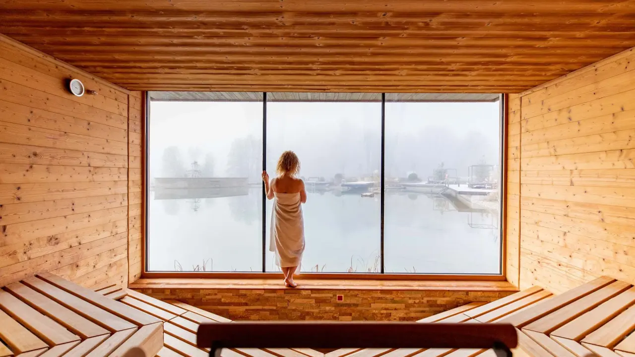Frau im Handtuch blickt aus Panoramafenster einer Holzsauna auf nebligen Naturbadesee im Pfalzblick Wald Spa Resort.
