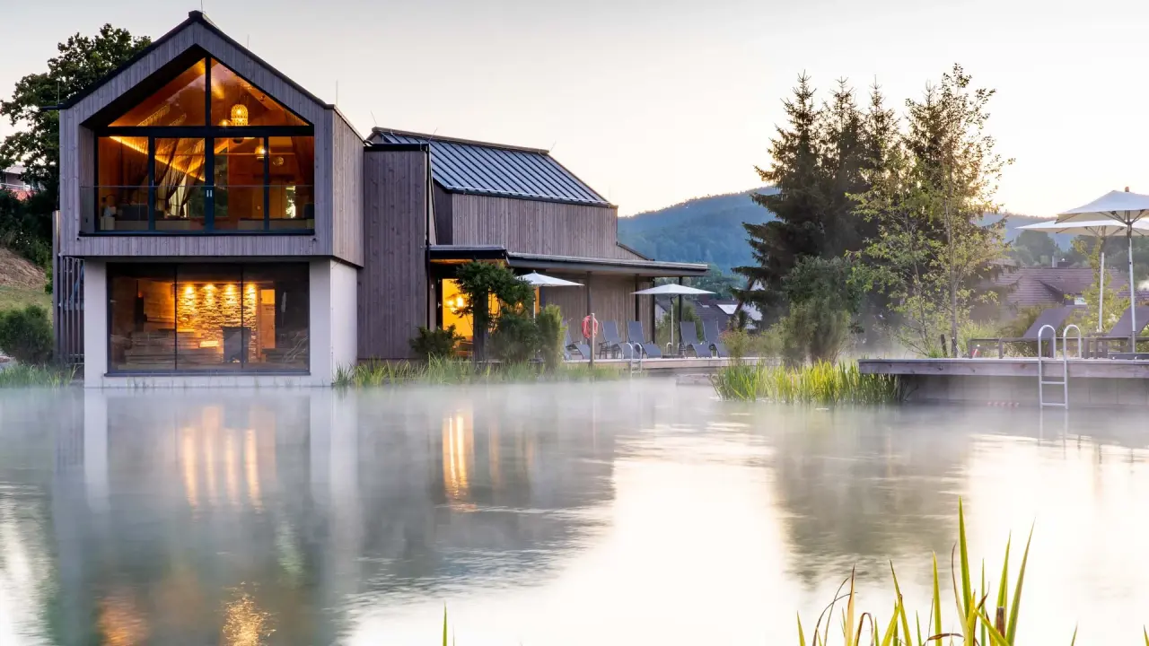 Malerischer Naturbadeteich mit leichtem Nebel und modernem Spa-Gebäude des Pfalzblick Wald Spa Resort.