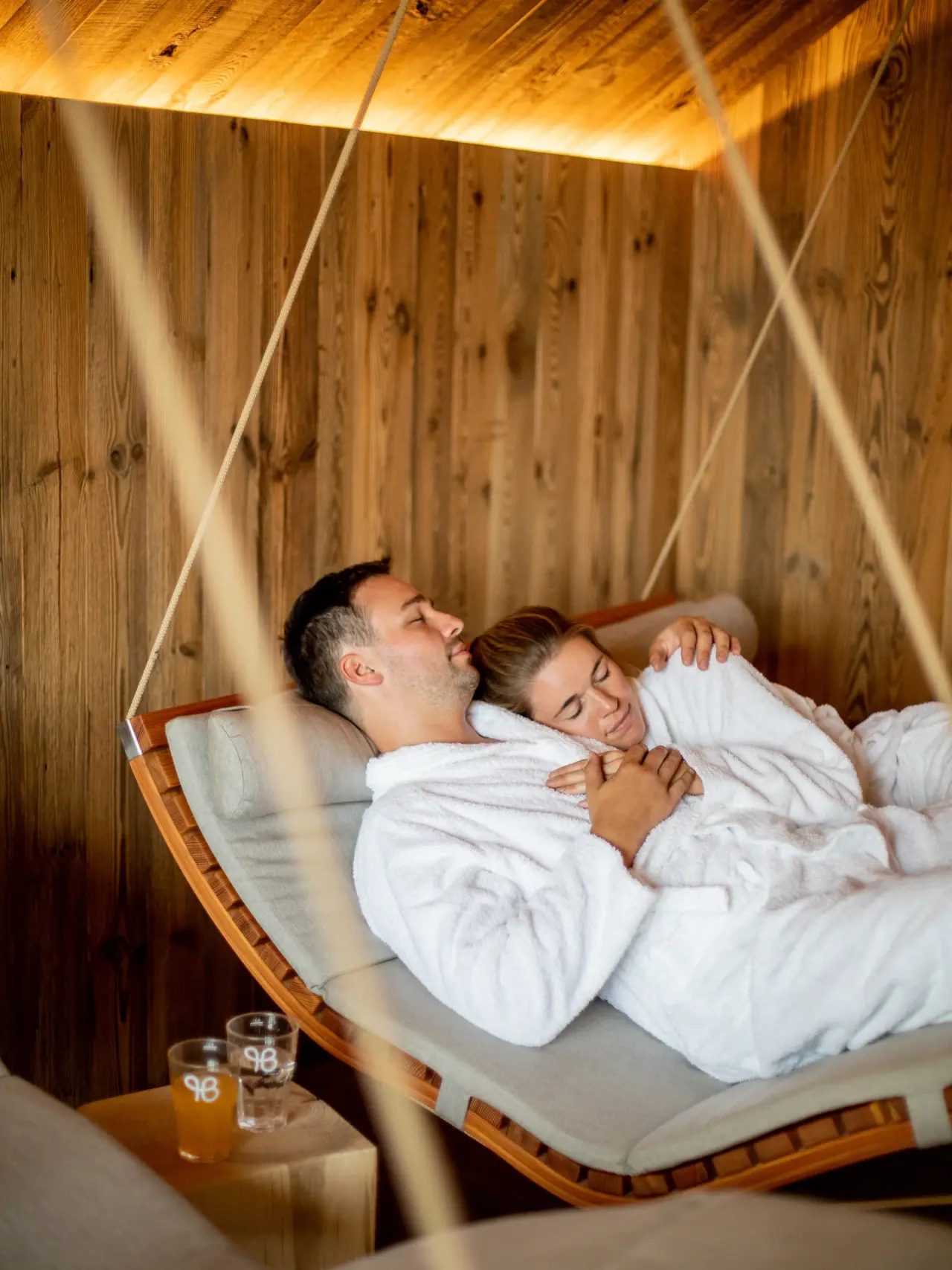 Paar entspannt in Bademänteln auf einer Schwebeliege im Wellnessbereich des Pfalzblick Wald Spa Resort.