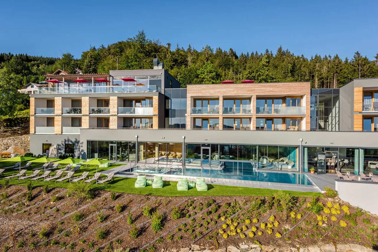 Modernes Wellnesshotel Hüttenhof mit großem Außenpool, Sonnenterrasse und grüner Berglandschaft unter blauem Himmel.