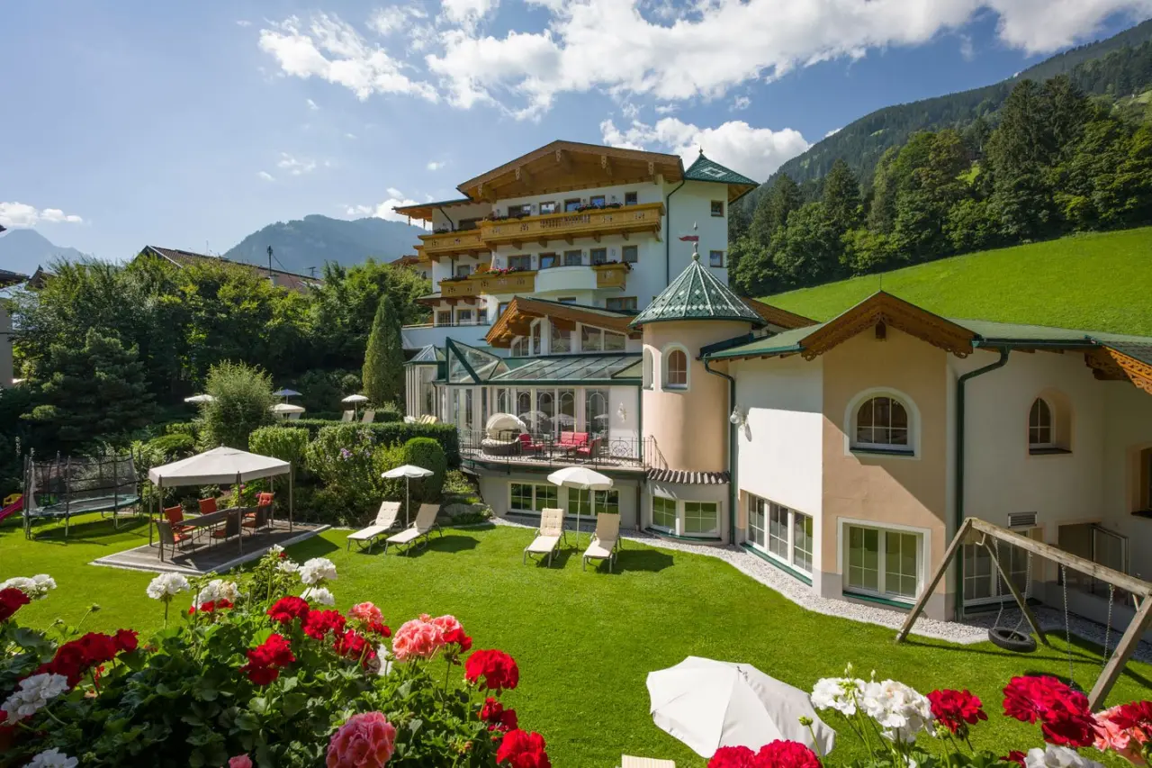 Ferienhotel Neue Post in den Bergen mit blühendem Garten, Sonnenterrasse und Alpenpanorama an einem sonnigen Tag.