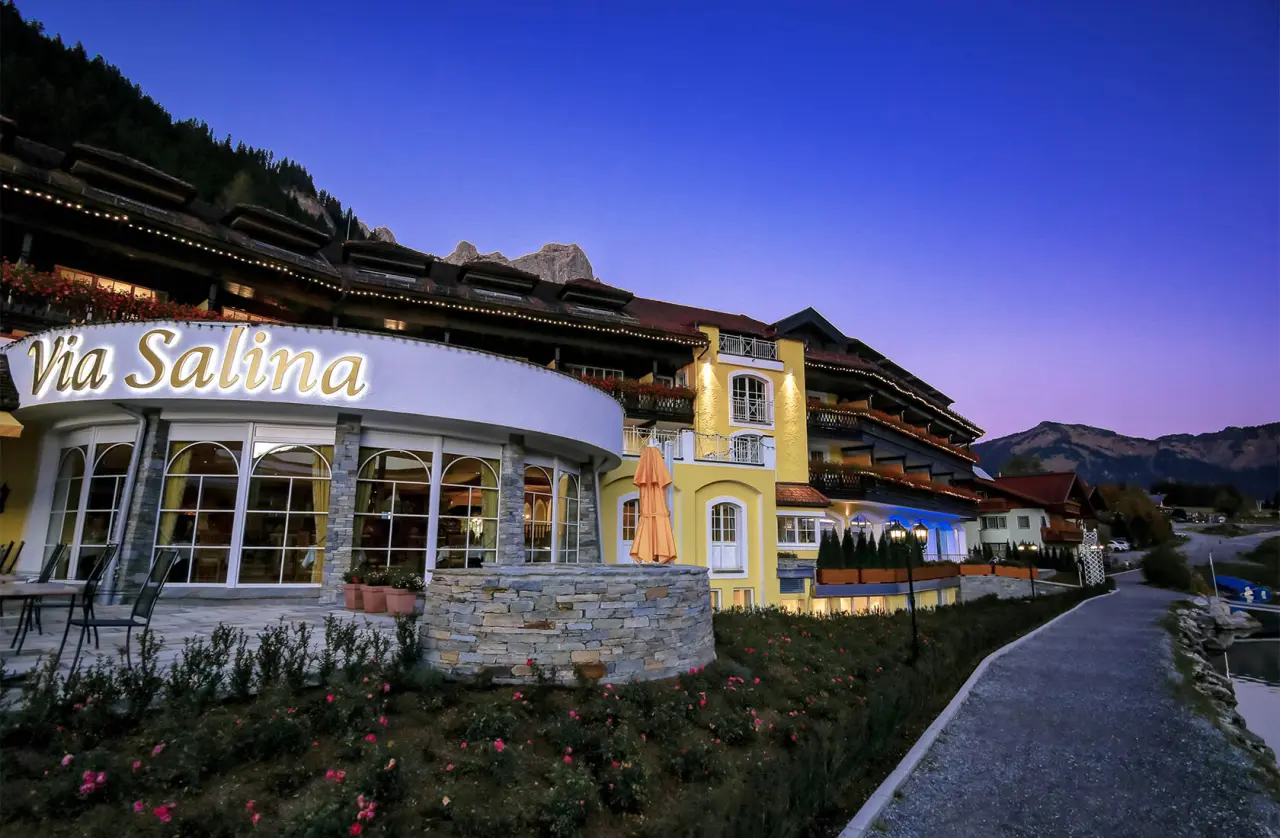 Beleuchtete Fassade des Hotel Via Salina bei Sonnenuntergang mit Bergkulisse und Seeweg.