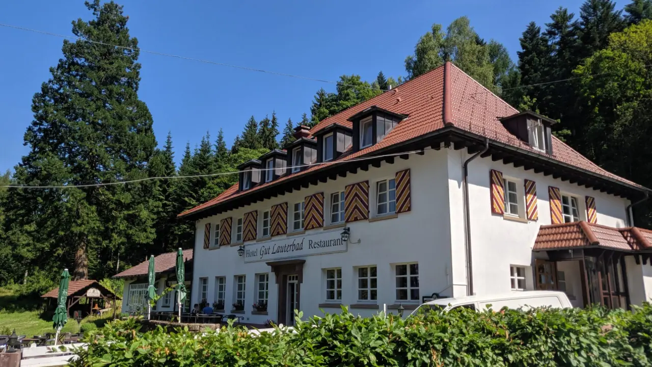 Aussenansicht des Hotel Gut Lauterbad Restaurant, umgeben von gruenen Baeumen unter blauem Himmel. Traditionelle Architektur.