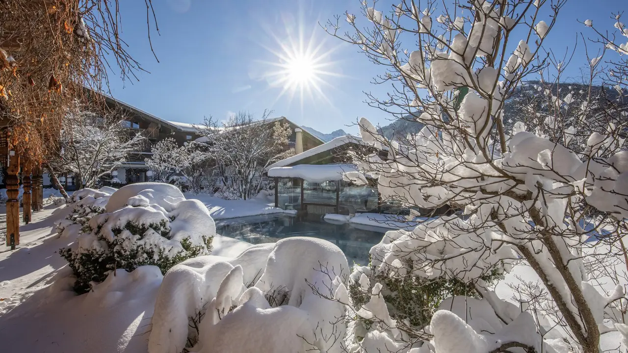 Beheizter Aussenpool im Schnee mit strahlender Wintersonne und dem Hotel Der Böglerhof - pure nature spa resort.