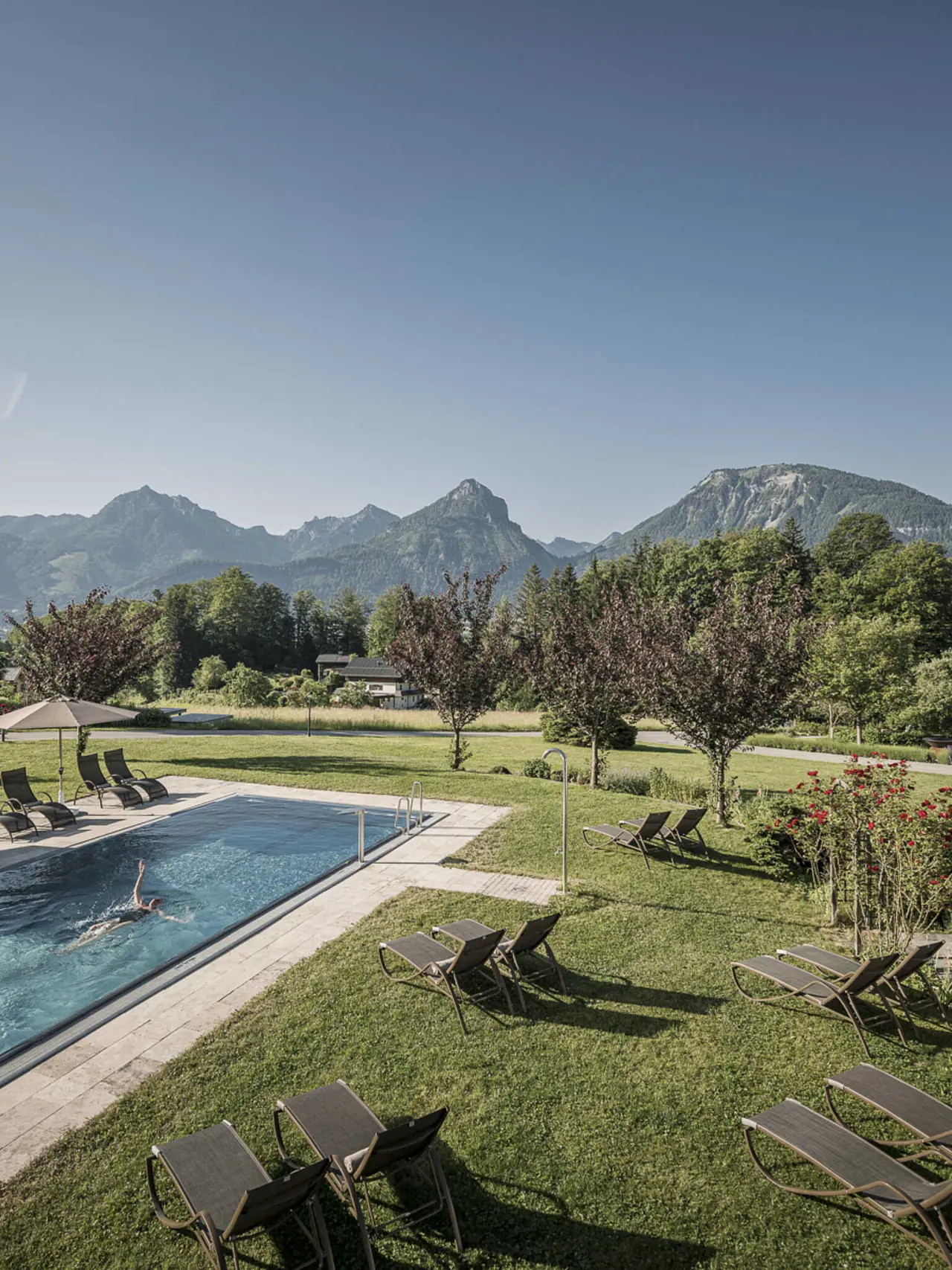 Aussenpool mit Liegewiese und beeindruckendem Bergpanorama im Hotel Bachmanngut. Eine Person schwimmt im klaren Wasser.