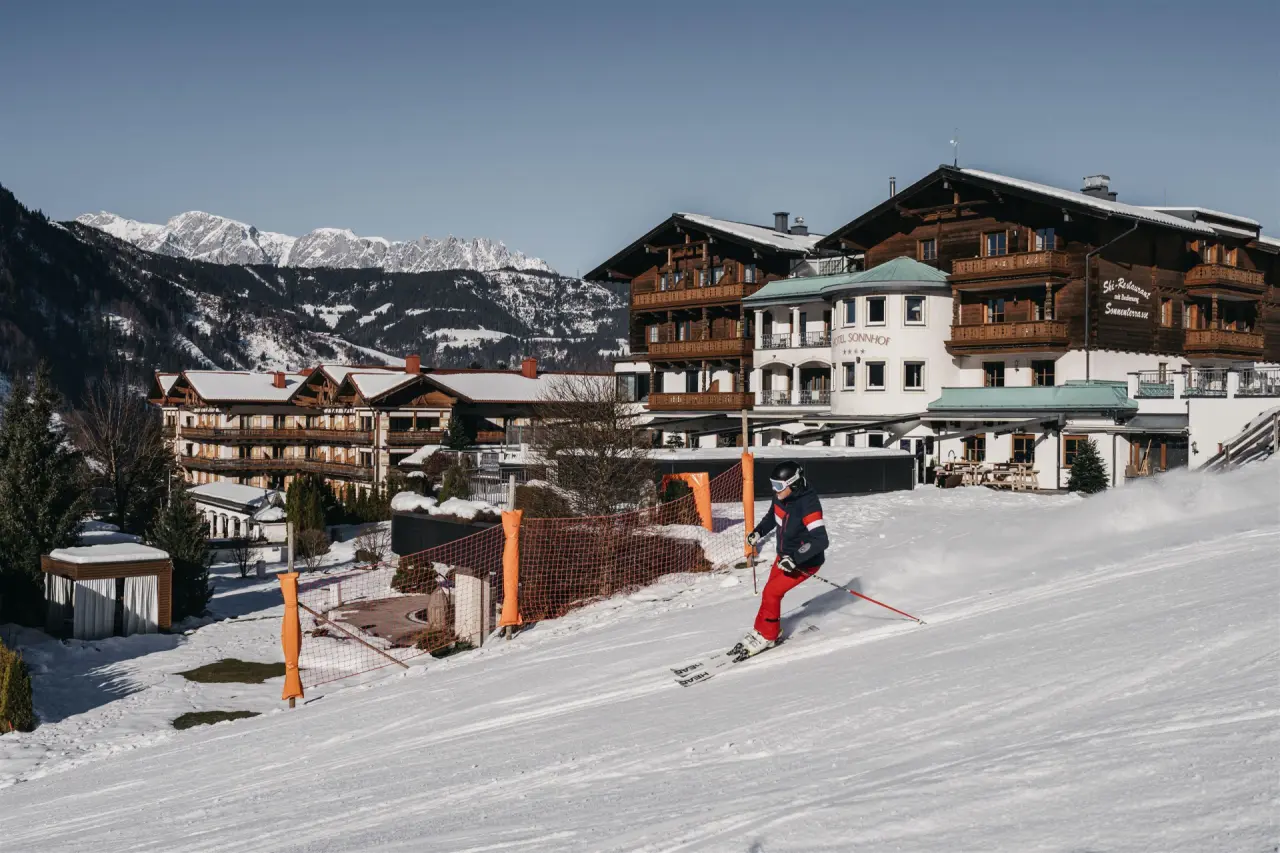 Skifahrer vor dem Hotel Sonnhof Alpendorf direkt an der Skipiste mit verschneiten Bergen im Hintergrund.