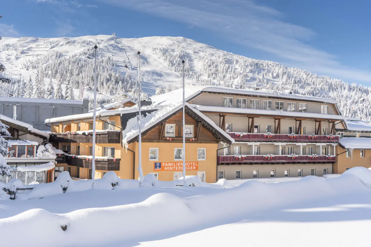 Familienhotel Hinteregger im Winter, umgeben von tiefem Schnee und verschneiten Bergen mit Skipisten unter blauem Himmel.
