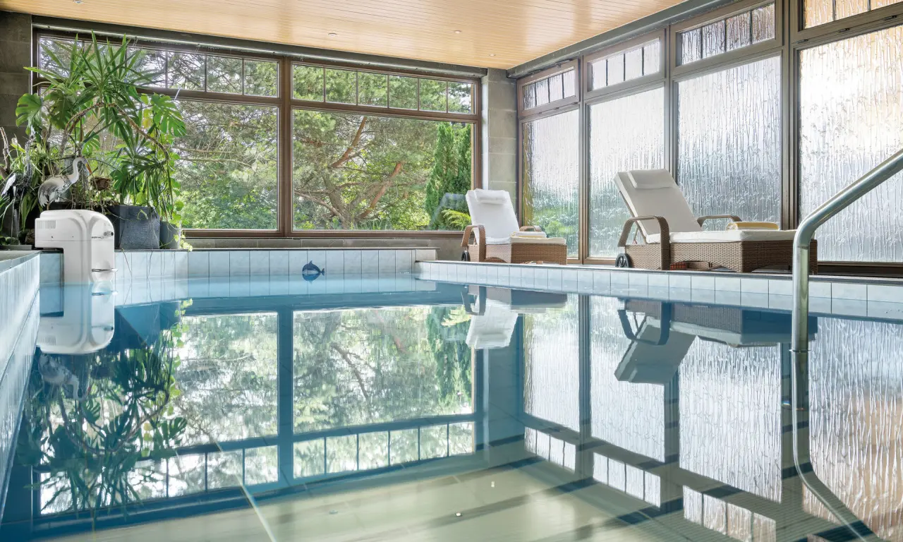 Indoor-Pool mit großen Fenstern und Blick ins Grüne im Schlossberghotel Oberhof. Entspannungsliegen am Beckenrand.