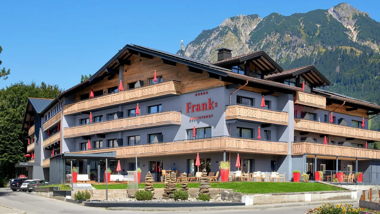 Aussenansicht des Hotel Franks in Oberstdorf mit traditioneller Architektur, Holzbalkonen und Bergpanorama unter blauem Himmel.