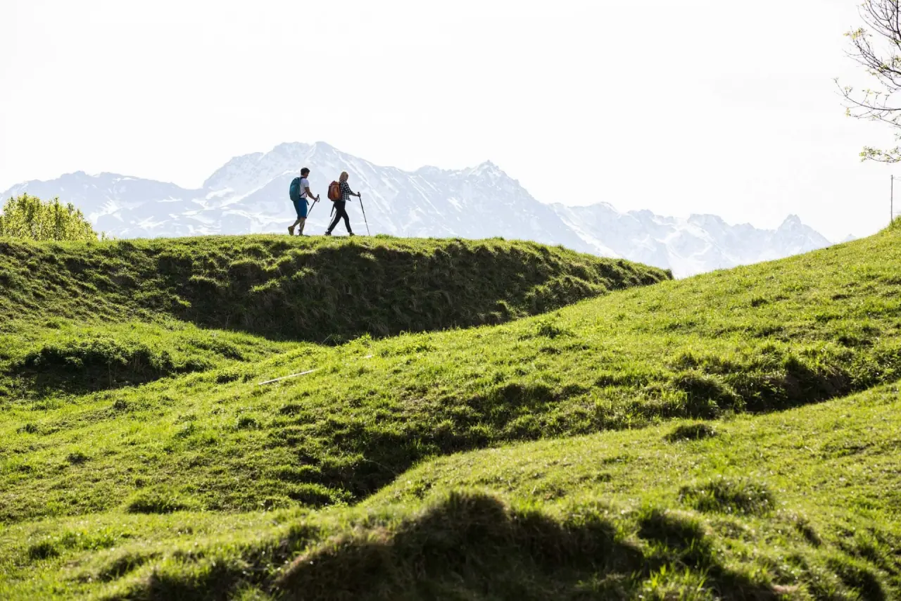 Zwei Wanderer mit Rucksäcken und Stöcken auf einem grünen Hügel mit Bergpanorama im Hintergrund, Hotel Jägerhof.