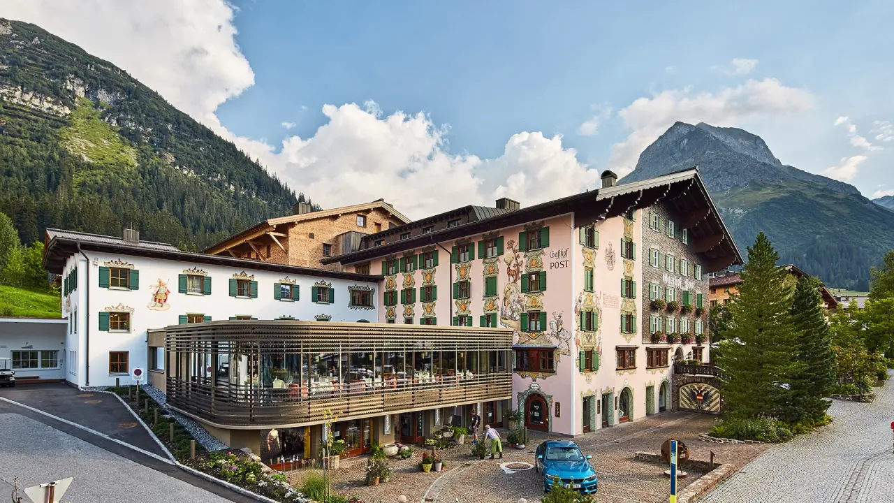 Traditionelles Hotel Post Lech mit bemalter Fassade und Glasanbau vor imposanter Bergkulisse in den Alpen.