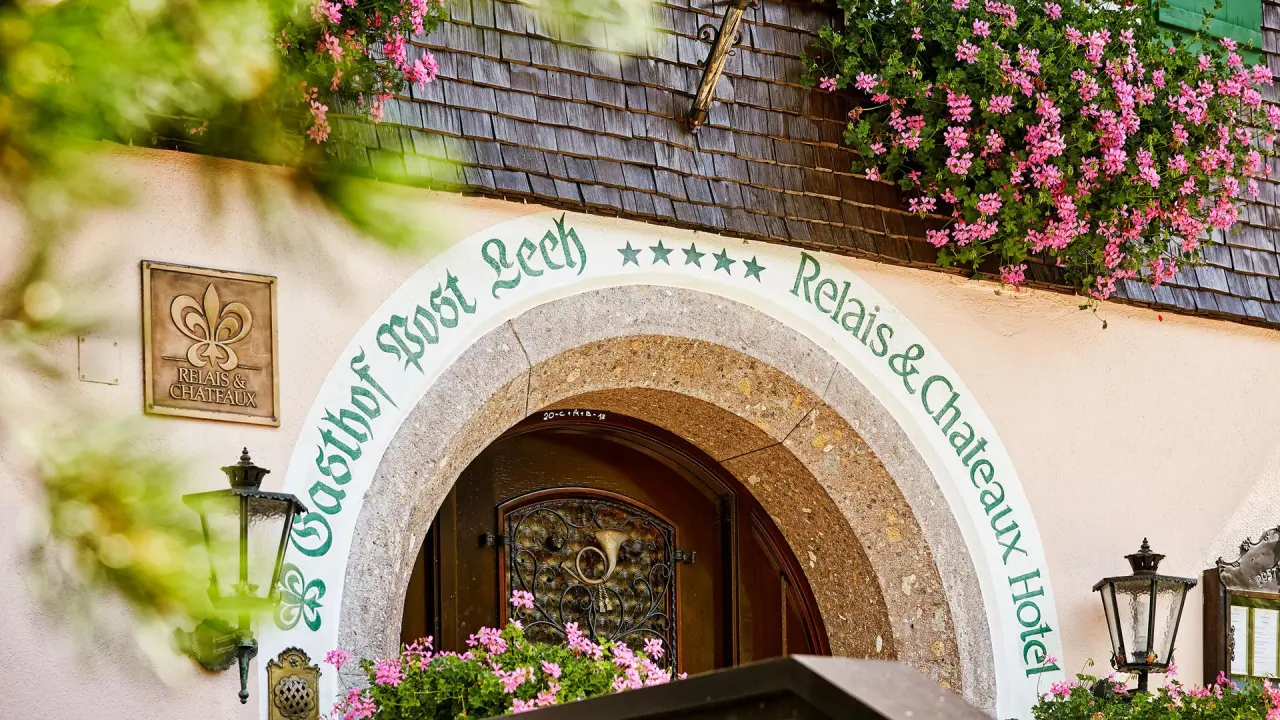 Traditioneller Eingang des Hotel Post Lech mit Blumen, Holzschindeldach und Relais & Chateaux Logo.