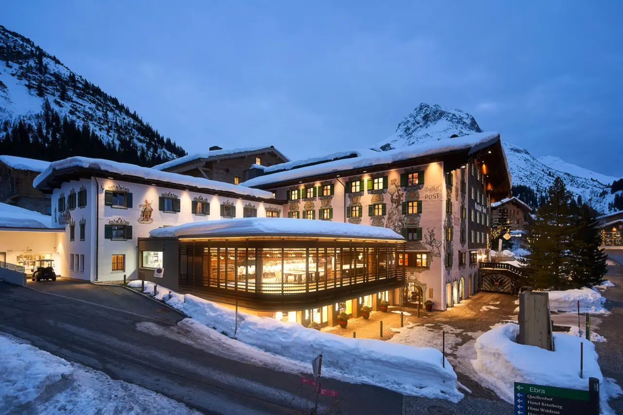 Abendansicht des Hotel Post Lech im Winter, umgeben von verschneiten Bergen. Warme Lichter beleuchten das traditionelle Alpenhotel.