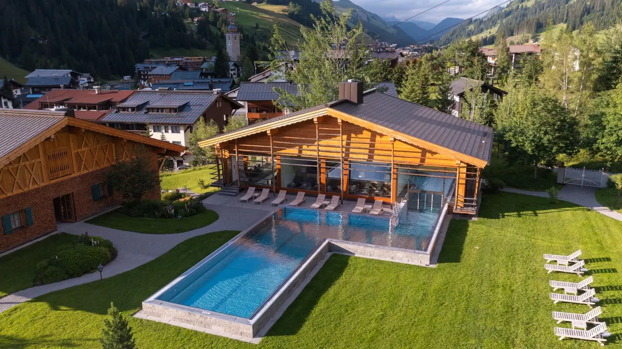 Aussenpool und Wellnessbereich mit Holzarchitektur im Hotel Post Lech, umgeben von grünen Wiesen und Alpenpanorama.