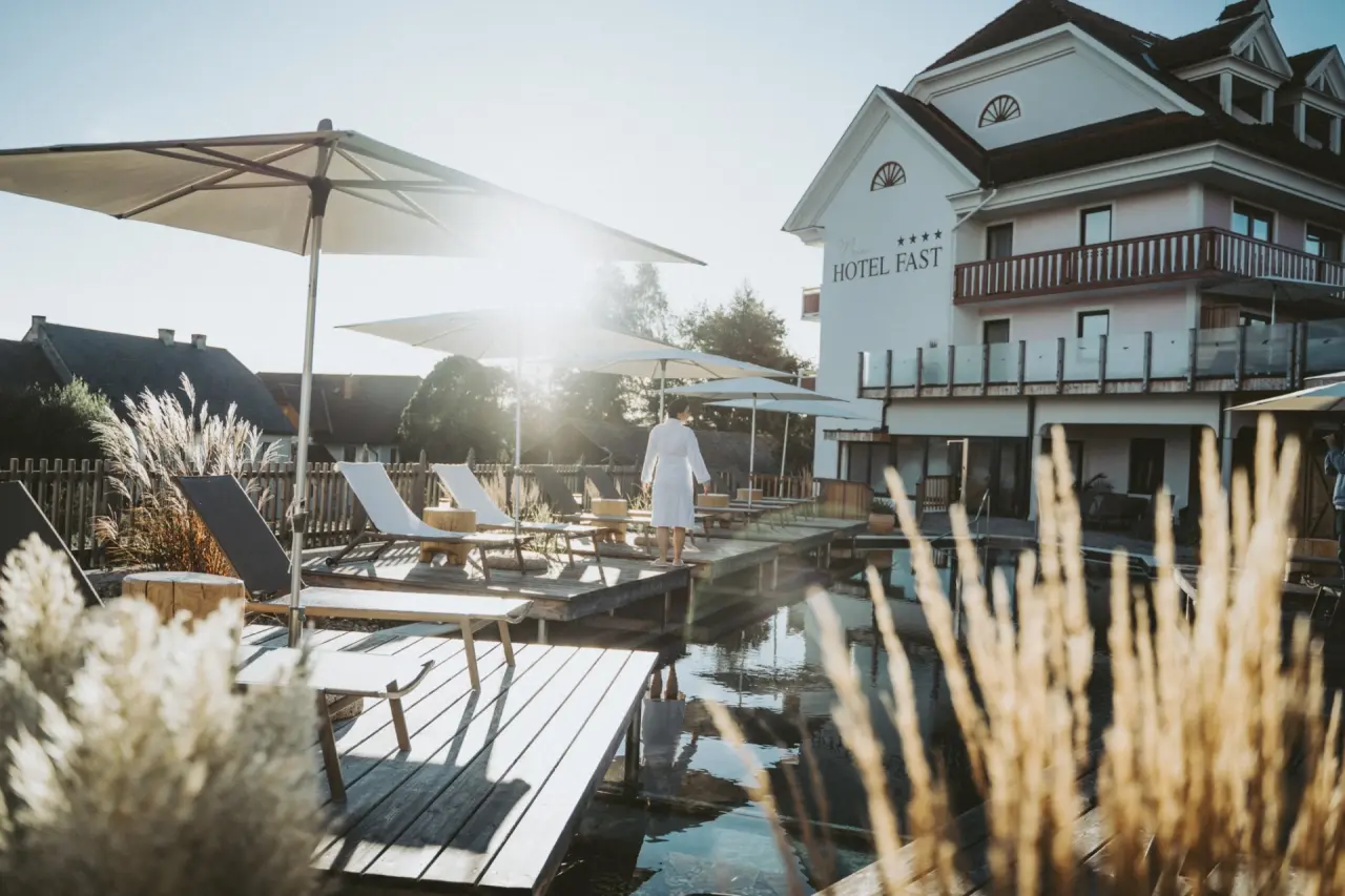 Natürlicher Badeteich mit Holzsteg, Liegen und Hotel Fast im warmen Abendlicht. Eine Person im Bademantel entspannt am Wasser.