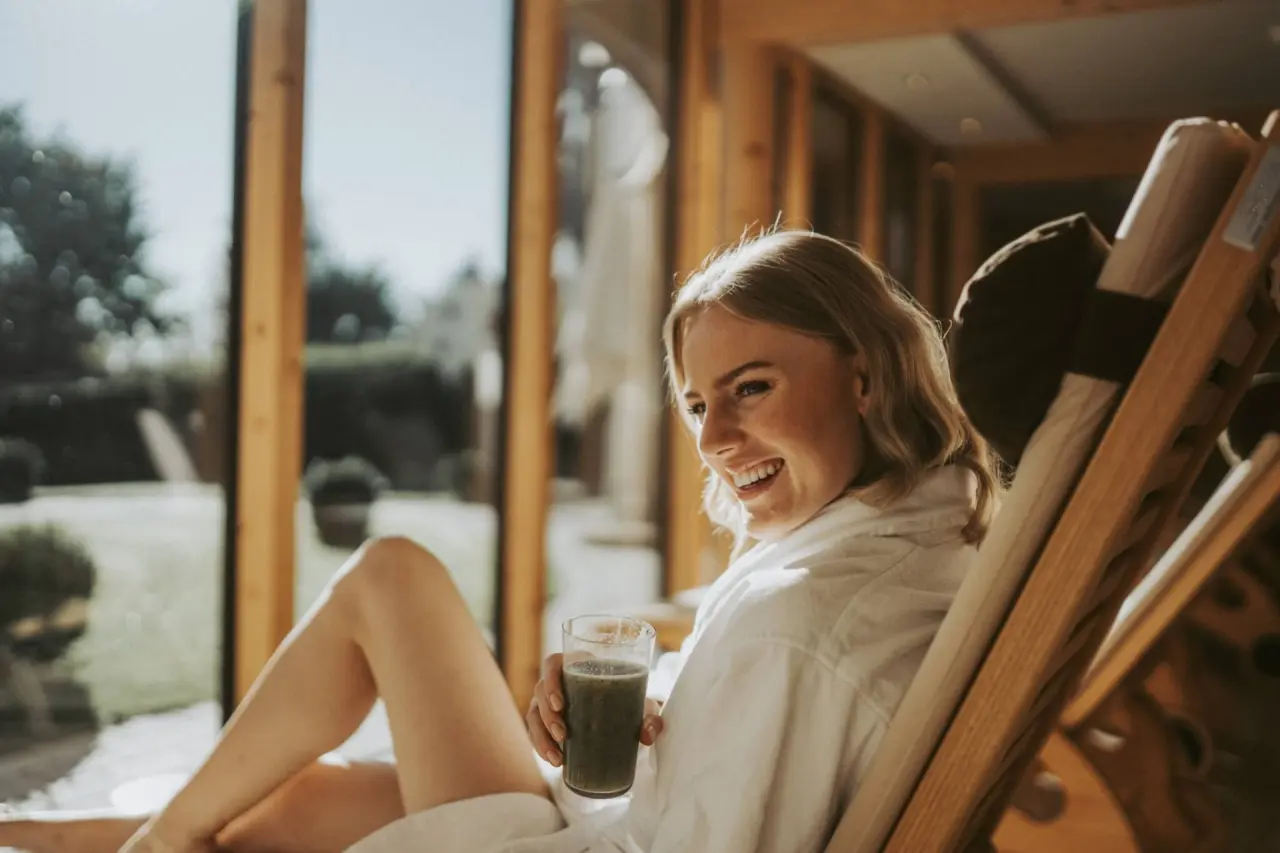 Lachende Frau im Bademantel entspannt mit gesundem Smoothie in der Wellness-Lounge von Mein Hotel Fast.