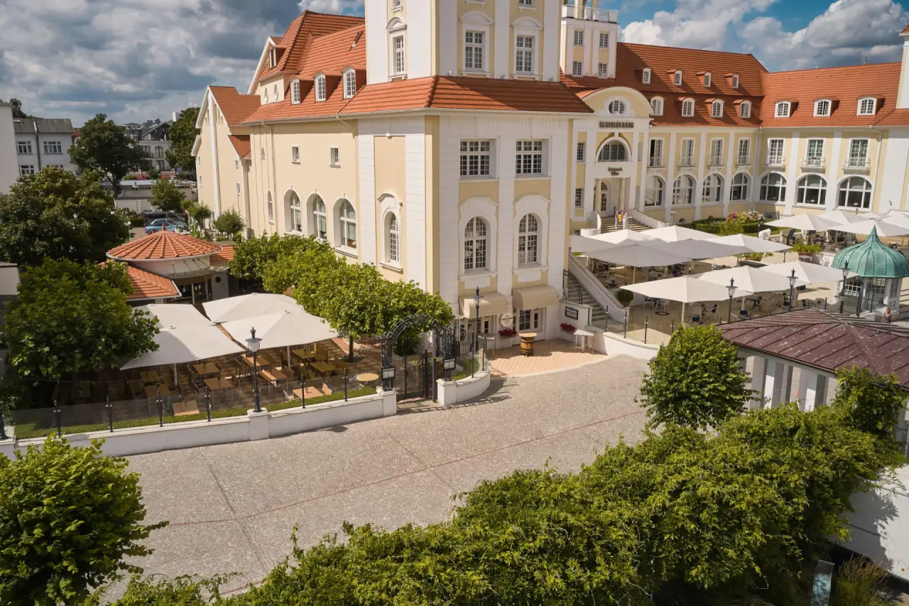 Luftaufnahme des eleganten Travel Charme Kurhaus Binz mit klassischer Architektur, Aussenrestaurants und grünen Bäumen.