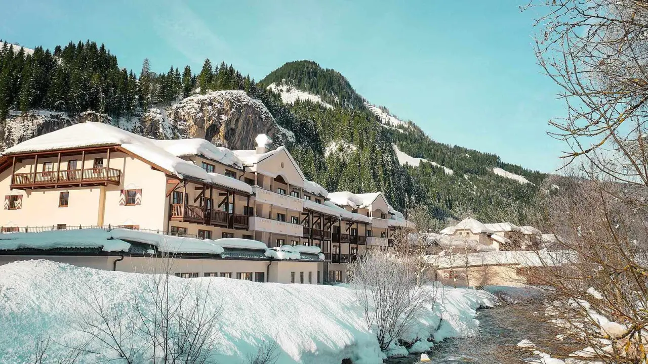 Winteransicht des ROBINSON Amadé Hotels, umgeben von verschneiten Bergen und Tannenwäldern, mit einem Bach im Vordergrund.