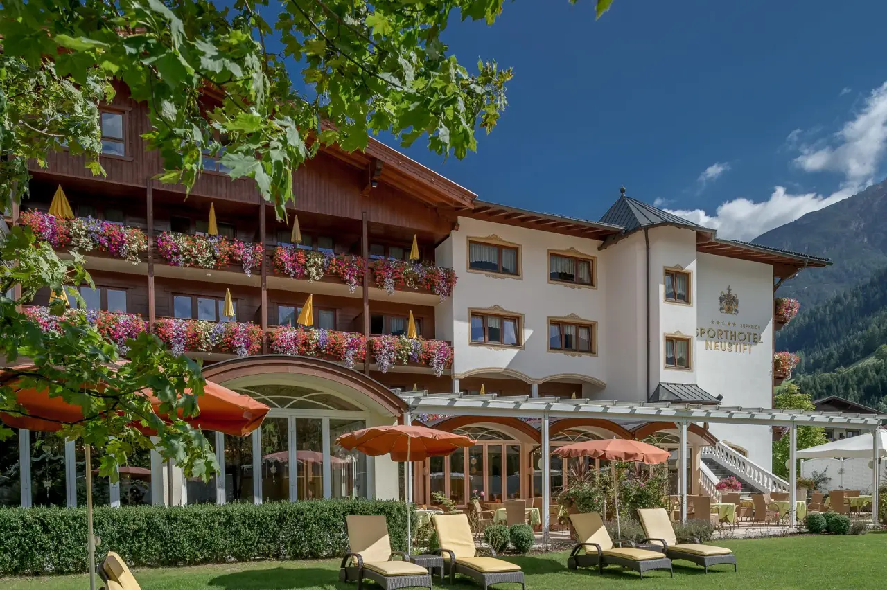 Sommeransicht des Sporthotel Neustift mit blühenden Balkonen, sonnigem Garten mit Liegestühlen und Bergpanorama.