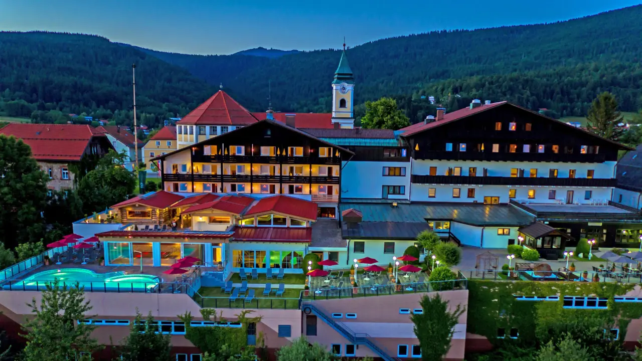 Abendliche Luftaufnahme des Wellnesshotel Hofbräuhaus mit beleuchtetem Aussenpool und Hotelanlage vor bewaldeten Bergen.