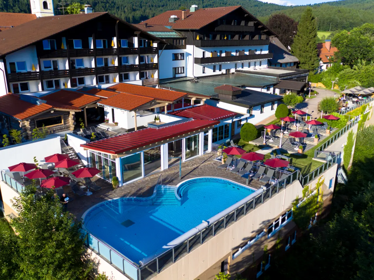 Luftaufnahme des Aussenpools mit Sonnenterrasse und roten Sonnenschirmen im Wellnesshotel Hofbräuhaus, umgeben von grüner Natur.