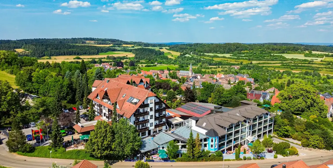 Luftaufnahme des Vital- und Wellnesshotel Albblick eingebettet in grüne Hügellandschaft mit Dorf im Hintergrund.