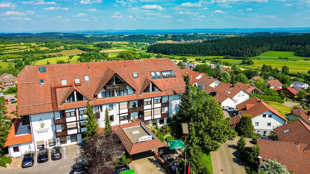 Luftaufnahme vom Vital- und Wellnesshotel Albblick mit rotem Dach, umgeben von grünen Wäldern und Feldern unter blauem Himmel.