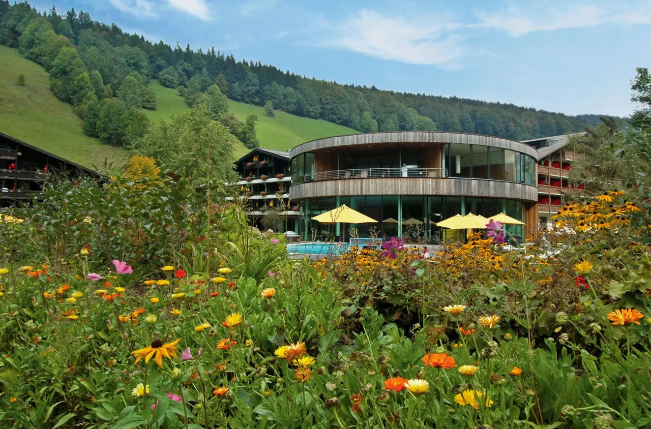 Aussenansicht des Gesundhotel Bad Reuthe mit modernem Rundbau, Aussenpool und blühendem Garten vor grünen Bergen.