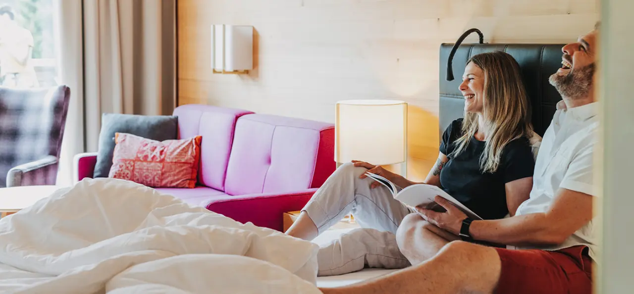 Gesundhotel Bad Reuthe****s in Reuthe – Glückliches Paar entspannt lesend im komfortablen Hotelzimmer | Wellnesshotel Bregenzerwald
