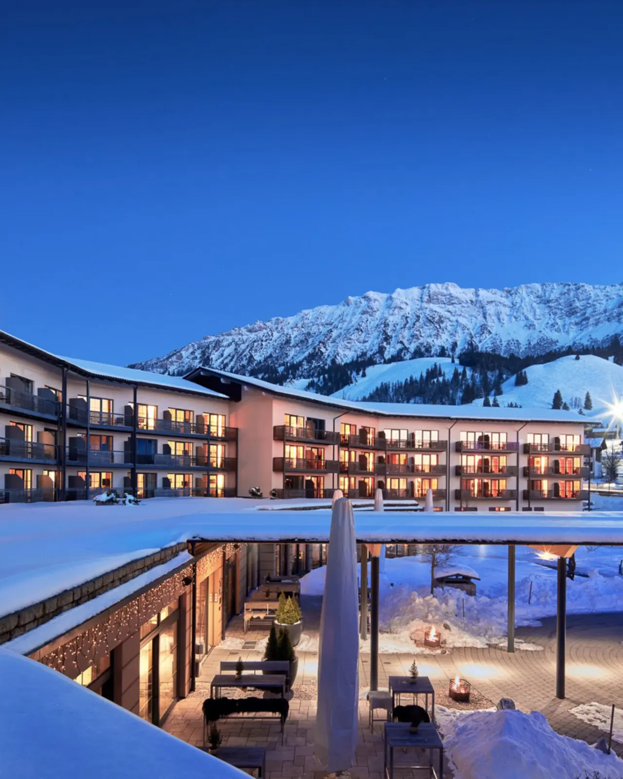 Panoramahotel Oberjoch im Winter bei Dämmerung. Beleuchtete Fassade mit Balkonen und verschneite Alpenkulisse.