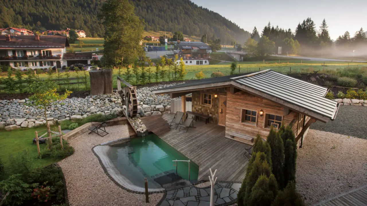 Idyllischer Naturpool mit Holzsauna und Wasserrad im Panoramahotel Oberjoch, umgeben von grüner Berglandschaft.