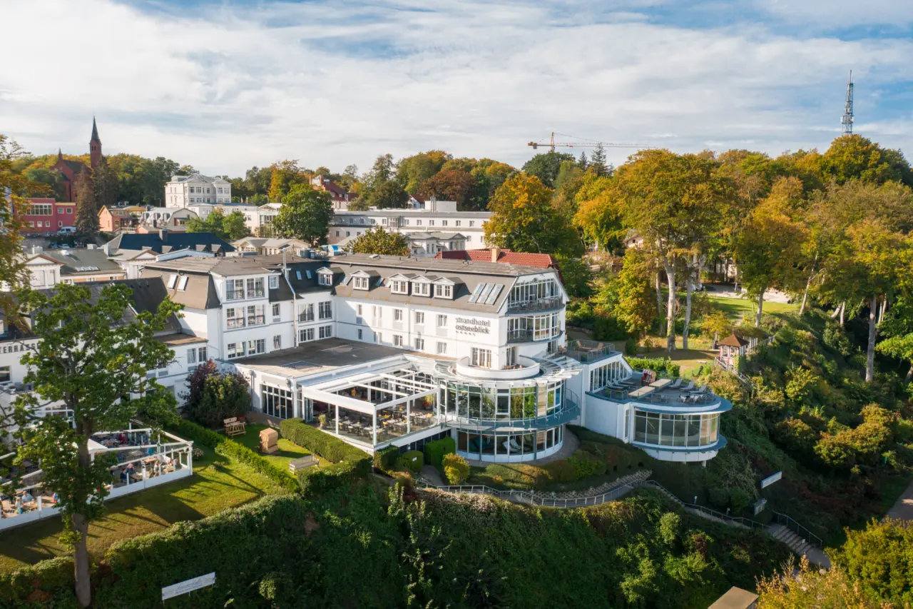 Luftaufnahme des Strandhotel Ostseeblick, ein elegantes weisses Hotelgebaeude umgeben von gruenen Baeumen unter blauem Himmel.