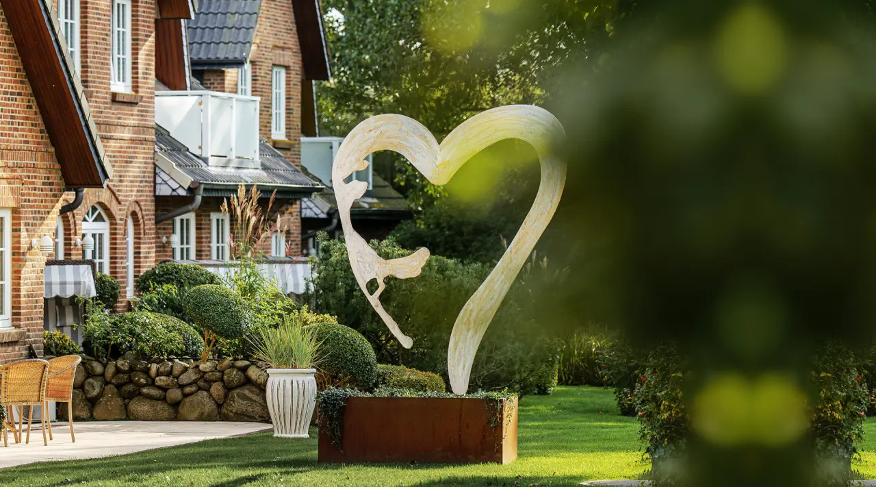 Gepflegter Hotelgarten mit Herzskulptur und traditioneller Backsteinfassade des Landhaus Stricker.