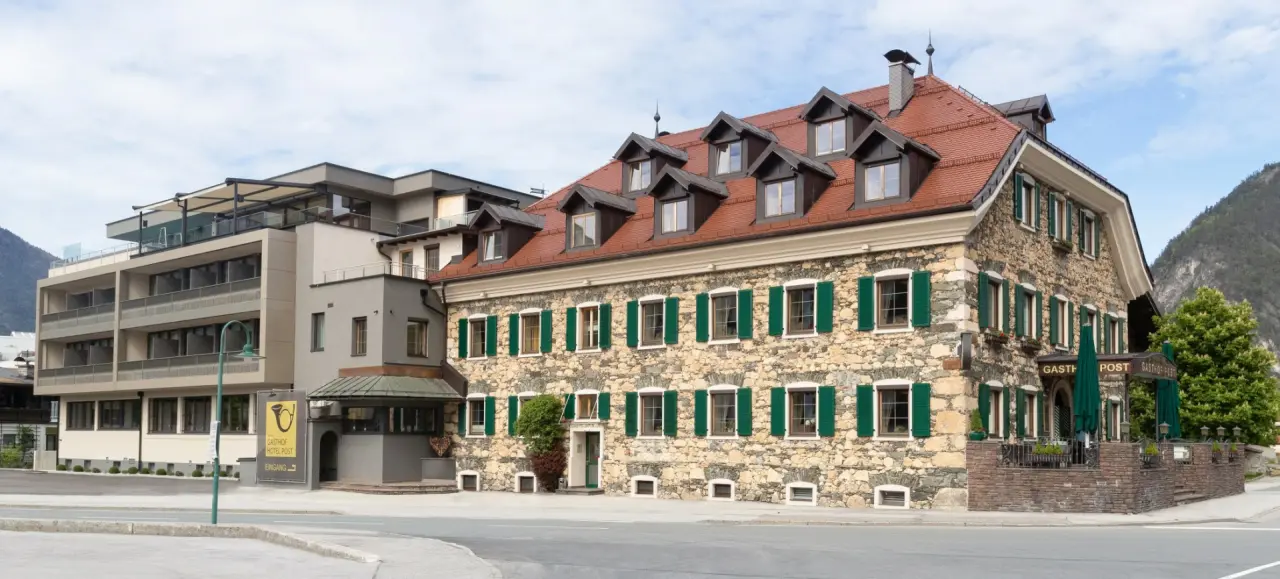 Aussenansicht des Gasthof Hotel Post mit traditionellem Steinbau und modernem Anbau vor Bergkulisse.