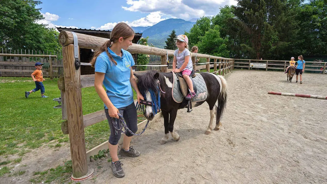 Kind auf Pony mit Reitlehrerin am Reitplatz des Hotel Glocknerhof vor einer malerischen Bergkulisse.
