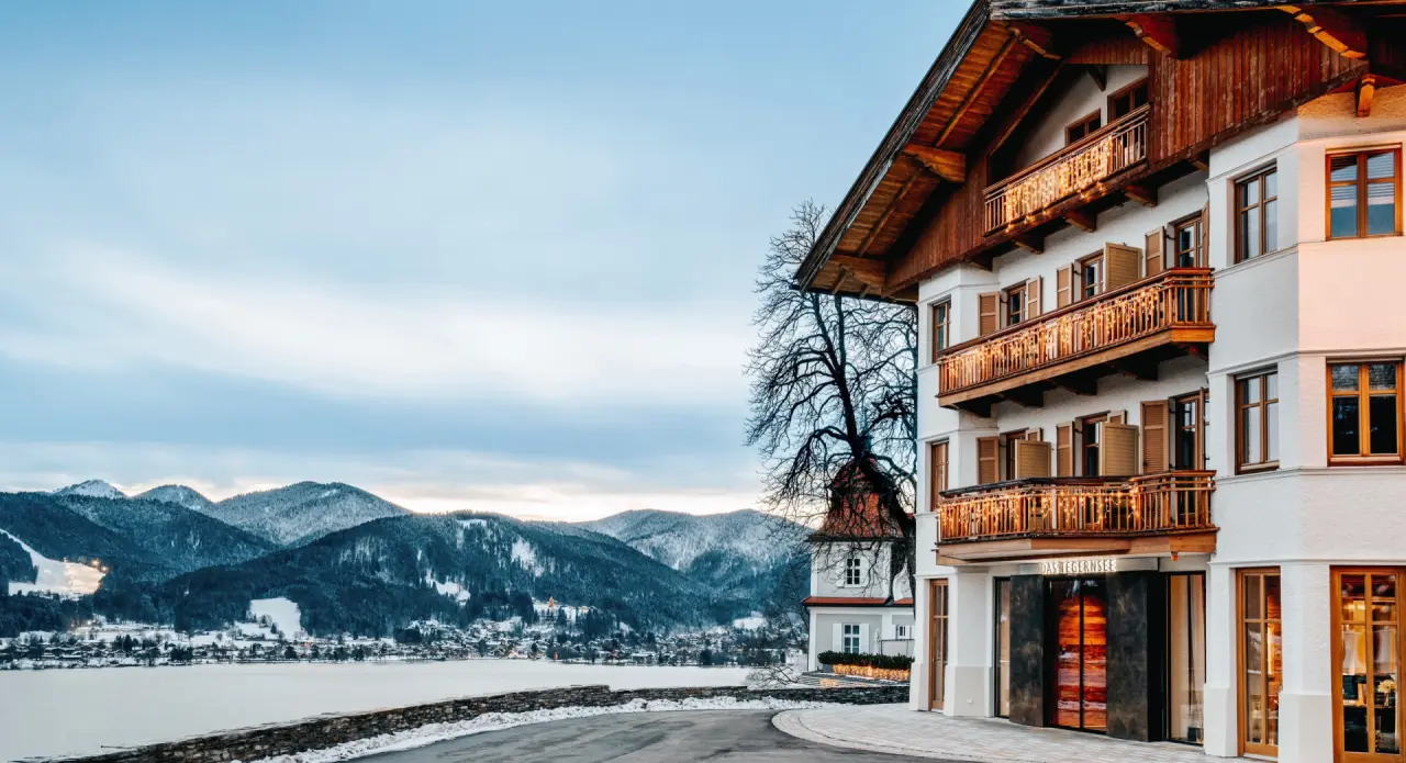 Winteransicht des Hotels DAS TEGERNSEE am verschneiten Tegernsee mit Alpenpanorama im Hintergrund.