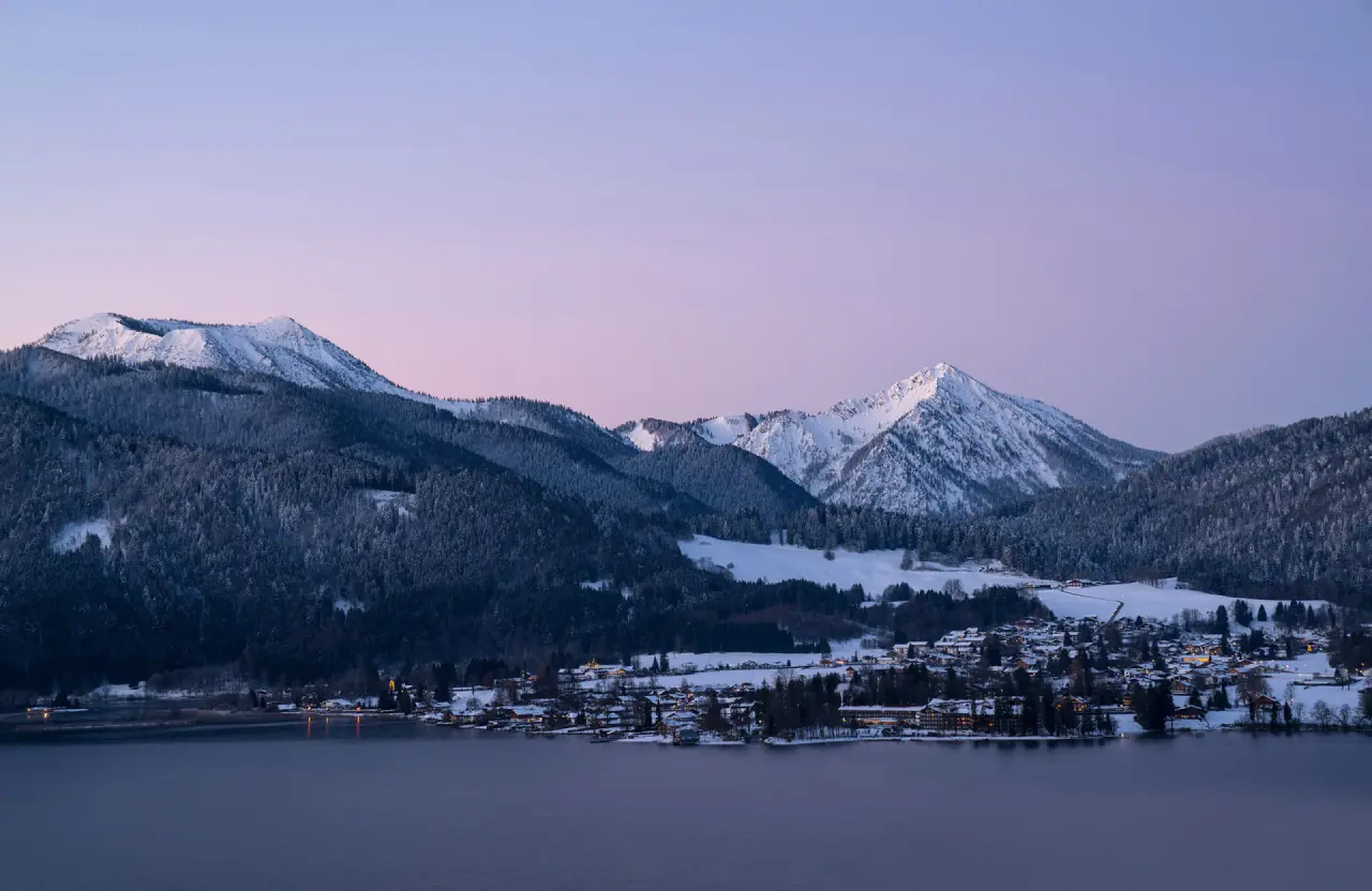Winterliches Panorama des Tegernsees mit verschneiten Bergen und dem beleuchteten Ort, inklusive Hotel DAS TEGERNSEE.