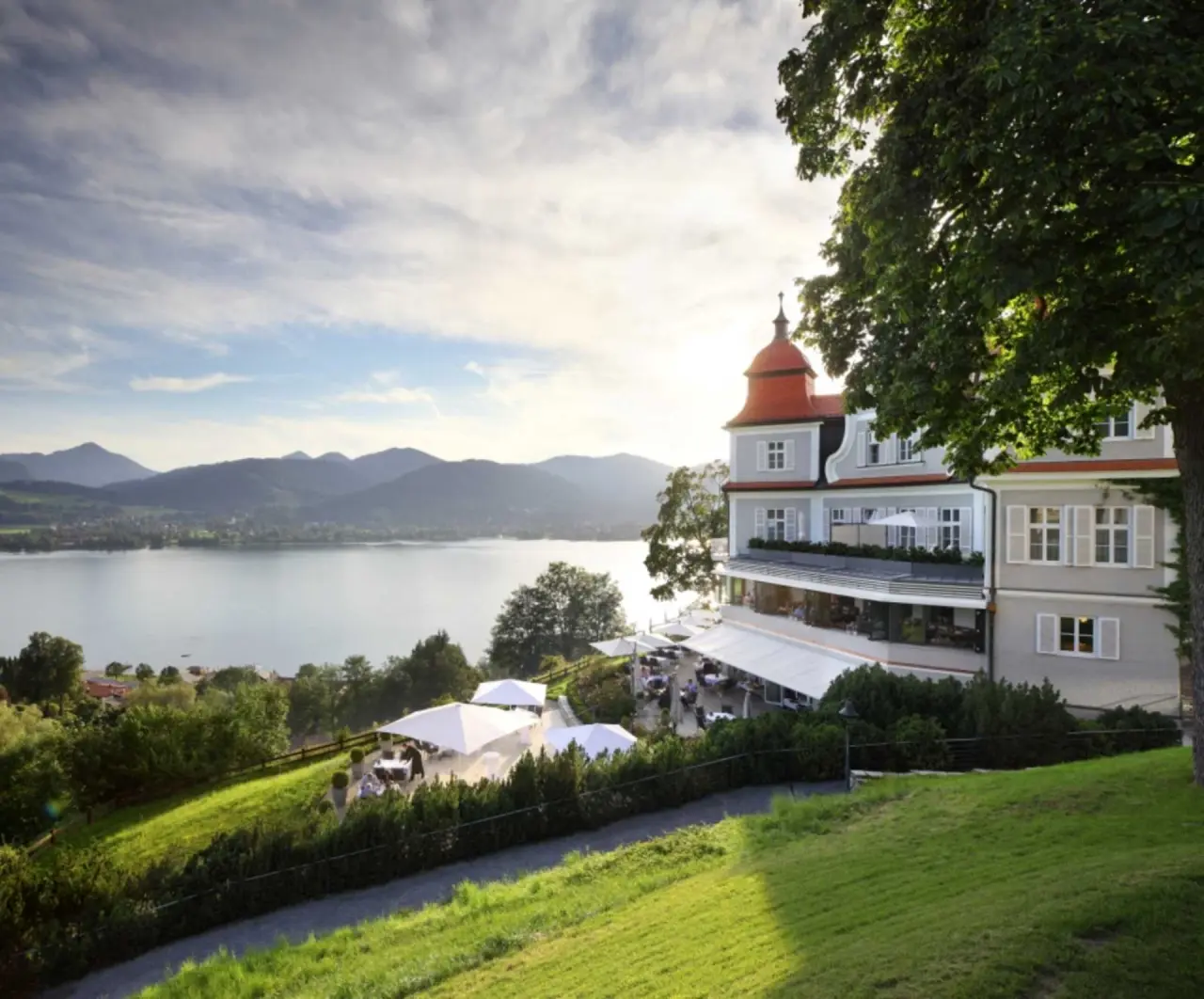 Panoramablick auf das Hotel DAS TEGERNSEE mit Terrasse, Tegernsee und Alpen im Hintergrund bei Sonnenuntergang.