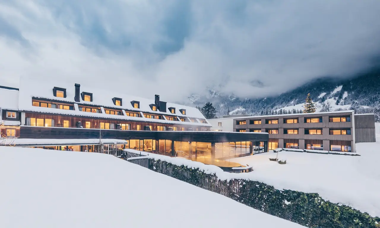 Winterliche Aussenansicht des TRAUBE BRAZ Alpen.Spa.Golf.Hotel mit verschneitem Dach und dampfendem Aussenpool vor Bergkulisse.