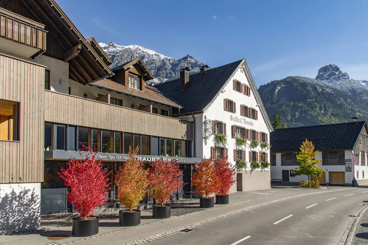Aussenansicht des TRAUBE BRAZ Alpen.Spa.Golf.Hotel mit traditioneller Architektur, bunten Herbstbäumen und schneebedeckten Alpen.